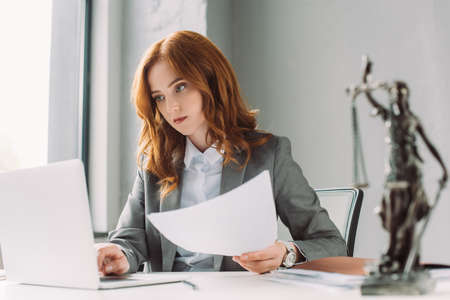 Redhead female lawyer with paper sheet looking at laptop at workplace with blurred themis figurine on foregroundの写真素材
