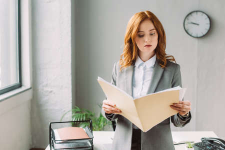 Serious businesswoman looking at folder with documents, while standing near workplace on blurred backgroundの写真素材
