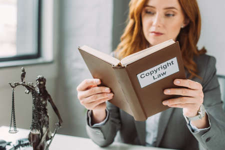 Concentrated redhead lawyer reading book with copyright law lettering near themis figurine with...