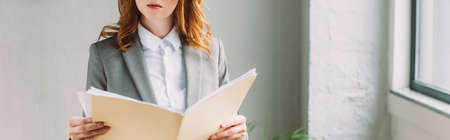 Cropped view of redhead businesswoman holding folder with paper sheets near window, bannerの写真素材