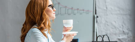 Side view of redhead businesswoman with coffee cup and saucer looking away with blurred flipchart on background, bannerの写真素材