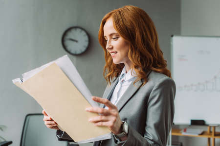 Smiling redhead businesswoman looking at folder with documents with blurred office on backgroundの写真素材