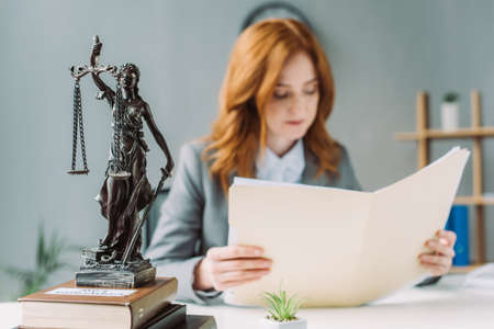 Female lawyer looking at folder, while sitting at workplace with themis figurine on pile of books on blurred backgroundの写真素材