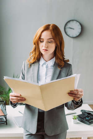 Serious businesswoman looking at folder with paper sheets, while standing near workplace on blurred backgroundの写真素材
