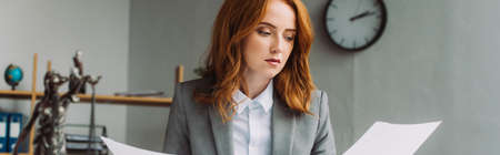 Redhead female lawyer looking at paper sheet with blurred themis figurine on foreground, banner