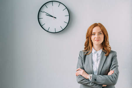 Confident businesswoman with crossed arms looking at camera near wall clock on grayの写真素材