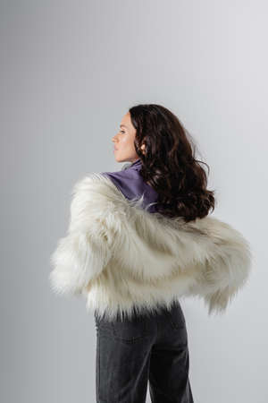 brunette young woman in stylish white faux fur jacket posing on gray background