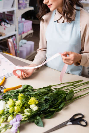 partial view of florist holding decorative ribbon while making bouquet from eustoma flowers in flower shop on blurred foregroundの写真素材