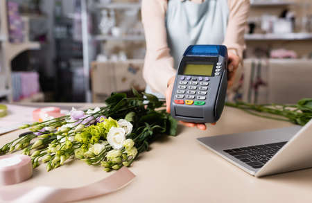 cropped view of florist holding payment terminal near laptop, eustoma flowers and decorative ribbon on blurred backgroundの写真素材