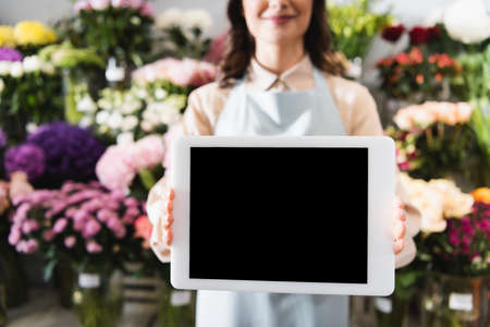 Cropped view of female florist showing digital tablet with blank screen with blurred range of flowers on backgroundの写真素材
