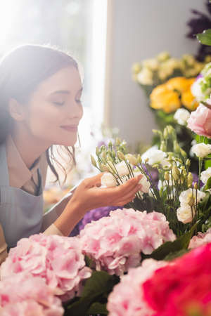 Happy florist with closed eyes smelling eustoma flowers near hydrangeas with blurred window on...