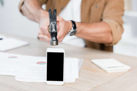 Cropped view of businessman holding hammer near smartphone with blank screen on workplace on blurred backgroundの写真素材