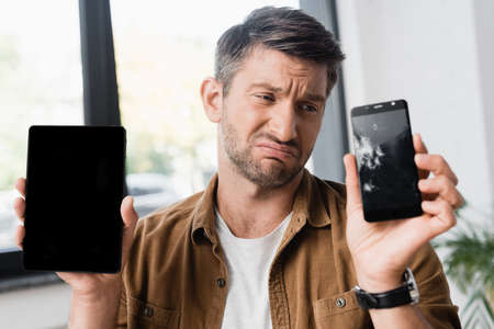 Sad businessman holding smartphone with blank screen while looking at smashed cellphone on blurred...