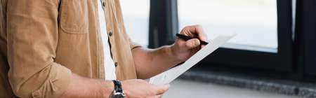 Cropped view of businessman standing and writing on document with blurred window on background, bannerの写真素材
