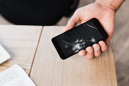 Cropped view of man holding smashed smartphone near table on blurred backgroundの写真素材