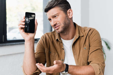 Upset businessman gesturing while looking at smashed smartphone with blurred window on background