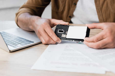 Cropped view of businessman putting battery in mobile phone at workplace with paperwork on blurred foregroundの写真素材
