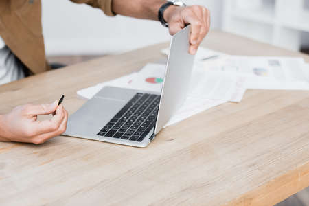 Cropped view of businessman with detail holding broken laptop at table with paperwork on blurred...