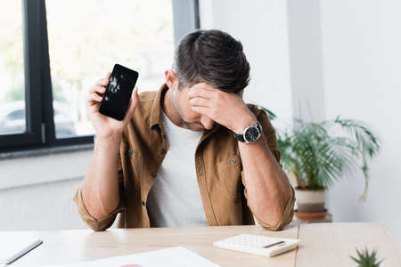 Disappointed businessman with smashed smartphone sitting at workplace on blurred backgroundの写真素材