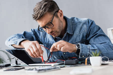 Focused repairman holding sensors of multimeter on broken keyboard of laptop at workplace on blurred...
