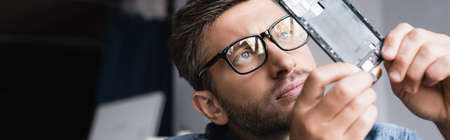 Focused repairman in eyeglasses looking at disassembled part of broken mobile phone on blurred...