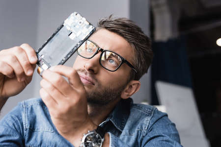 Focused repairman in eyeglasses looking at disassembled component of mobile phone on blurred...