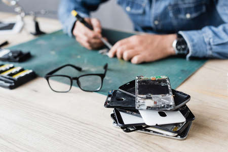 Close up view of pile with broken mobile phones near eyeglasses on workplace with blurred repairman...