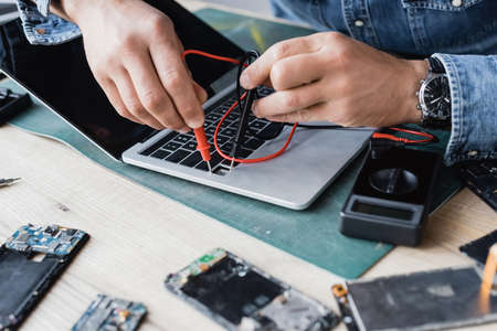 Cropped view of repairman holding sensors of multimeter on broken keyboard of laptop with blurred...