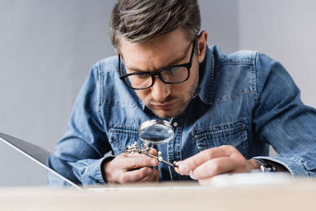 Focused repairman in eyeglasses with magnifier looking through at broken laptop with blurred...