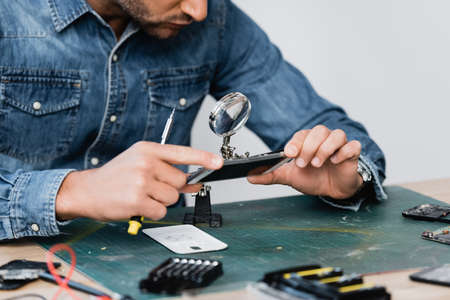 Cropped view of repairman looking through magnifier at disassembled part of cellphone at workplace on blurred foregroundの写真素材