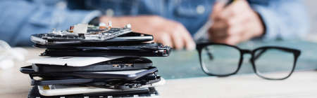 Close up view of pile of broken mobile phones near eyeglasses on workplace with blurred repairman on background, bannerの写真素材