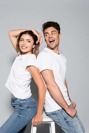 smiling young couple in white t-shirts and jeans posing back to back isolated on grey background