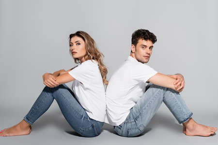 young couple in white t-shirts and jeans posing back to back isolated on grey background