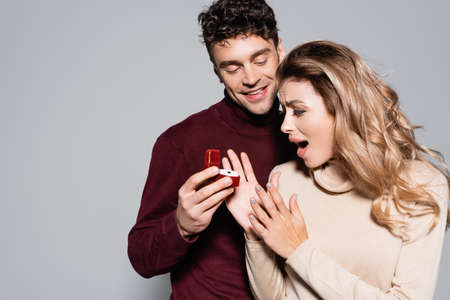 casual young man doing marriage proposal to shocked woman isolated on grey background