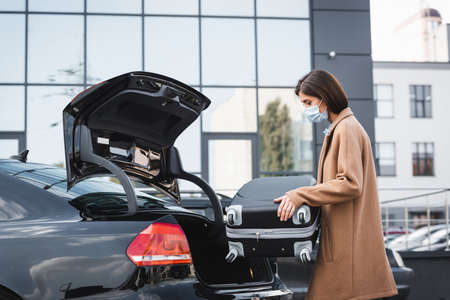 woman in trench coat and medical mask putting suitcase in car trunk