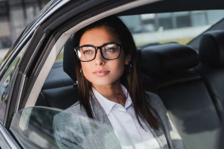 young businesswoman in eyeglasses looking out from open window while riding in car on blurred...
