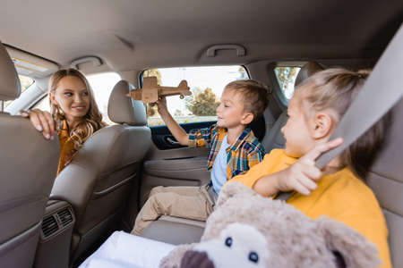 Smiling boy holding toy plane near mother and sister on blurred foreground in car