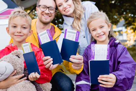 Smiling kids holding passports with air tickets near parents and car on blurred background outdoors
