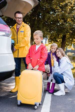 Smiling boy in autumn outfit holding suitcase near family and car outdoors