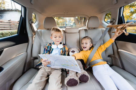 Cheerful kids with toys and map sitting on back seat of car