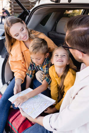 Smiling girl looking at father near family with map in trunk of car