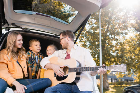 Smiling father playing acoustic guitar near wife and kids in car trunkの写真素材