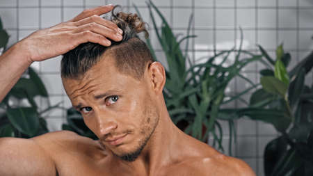 shirtless young man looking at camera adjusting hair near green plants on blurred backgroundの写真素材