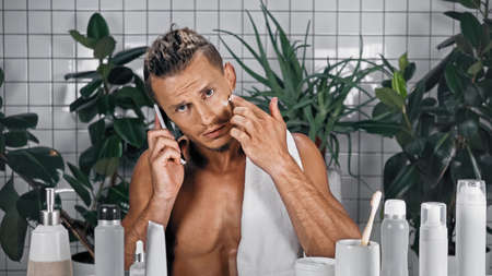 bearded man talking on smartphone and applying face cream in bathroom near plants on blurred backgroundの写真素材