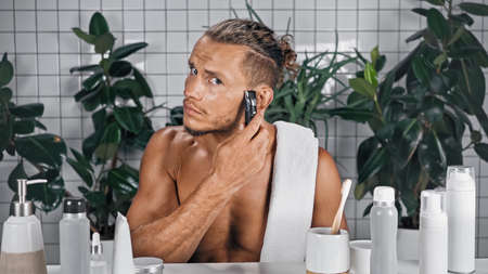 shirtless man using trimmer in bathroom near bottles and plants on blurred backgroundの写真素材