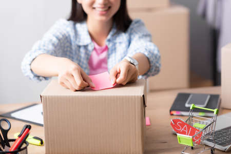 Cropped view of volunteer pasting sticker on carton box near shopping cart while sitting at desk on...