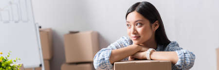 Positive asian volunteer looking away while leaning on carton box in charity center on blurred...