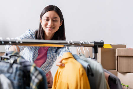 Smiling asian volunteer standing near clothes on hanging rack on blurred foreground