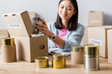 Smiling asian volunteer packing canned food in box with donations lettering in charity center