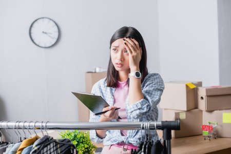 Upset asian volunteer holding clipboard with pen near hanging rack with clothes on blurred foreground in charity centerの写真素材